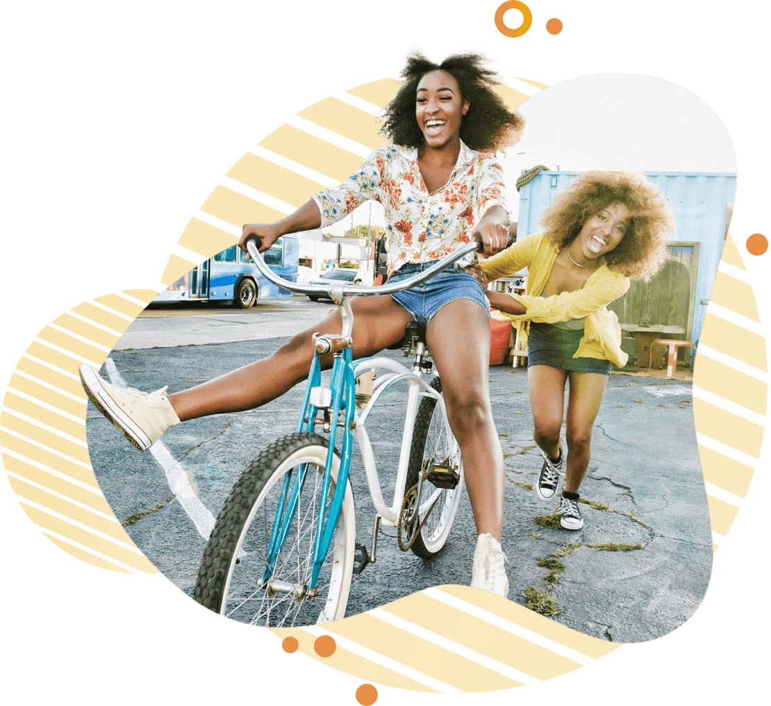 Two girls with smiling faces, one riding the bicycle while the other pushes it